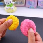 Two hands gently squeezing vibrant yellow and pink textured squishy balls made from balloons filled with micro styrofoam beads against a dark crafting mat.
