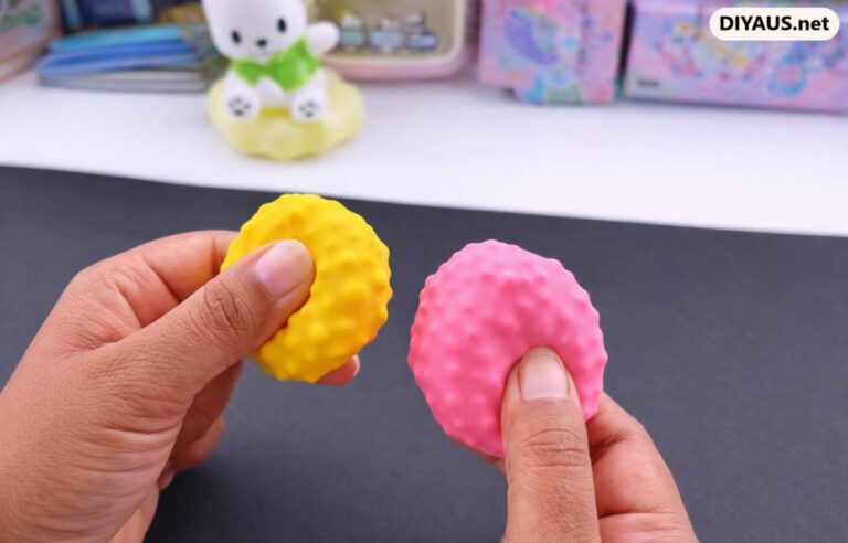 Two hands gently squeezing vibrant yellow and pink textured squishy balls made from balloons filled with micro styrofoam beads against a dark crafting mat.